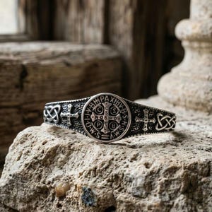 May include: A silver-toned metal cuff bracelet featuring a central circular Saint Benedict medal with embossed Latin lettering and a cross. The band is decorated with additional small crosses and intricate Celtic knotwork patterns on both sides.