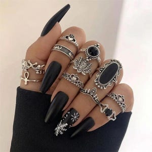 May include: A collection of silver-toned rings worn on fingers with long black nails, including designs featuring an ankh, a lotus flower, a large oval black stone, crowns, and intricate filigree patterns with small black accents.