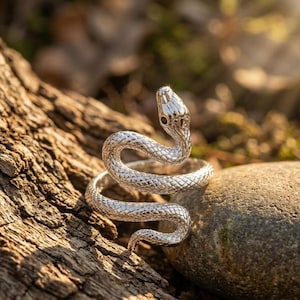 Op de afbeelding: Een zilverkleurige ring in de vorm van een opgerolde slang met een textuur die op schubben lijkt. De slangenkop bevindt zich aan de bovenzijde met kleine donkere ogen, terwijl het lichaam zich rondbuigt om de ringband in een spiraal te vormen.