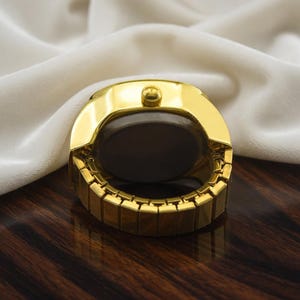 May include: A gold-toned ring watch with a dark face and a rectangular band. The watch has a rounded top with a small button and is set against a white fabric background.