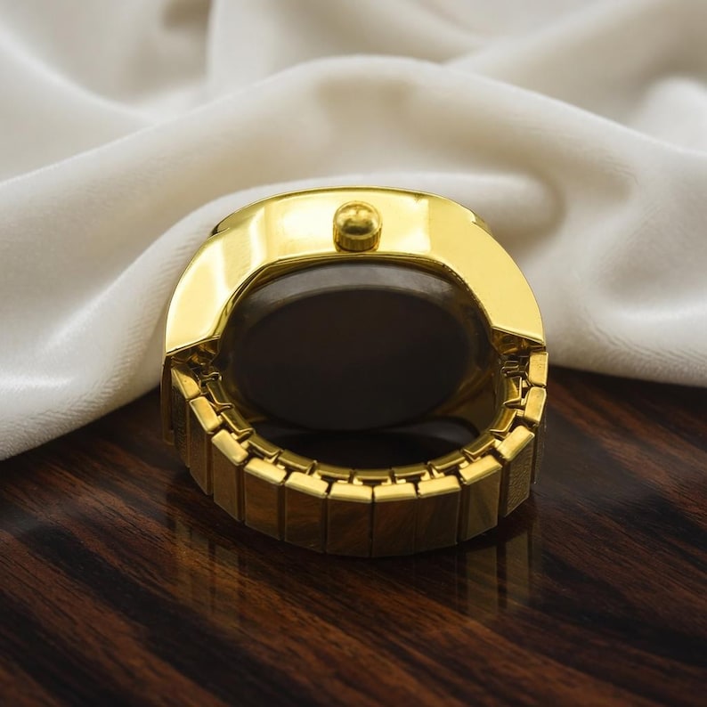 May include: A gold-toned ring watch with a dark face and a rectangular band. The watch has a rounded top with a small button and is set against a white fabric background.