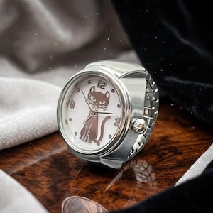 May include: A silver ring watch with a white face featuring a brown cat design. The watch has a round face with black numerals at the 12, 3, and 9 o'clock positions. The watch band is made of silver links.