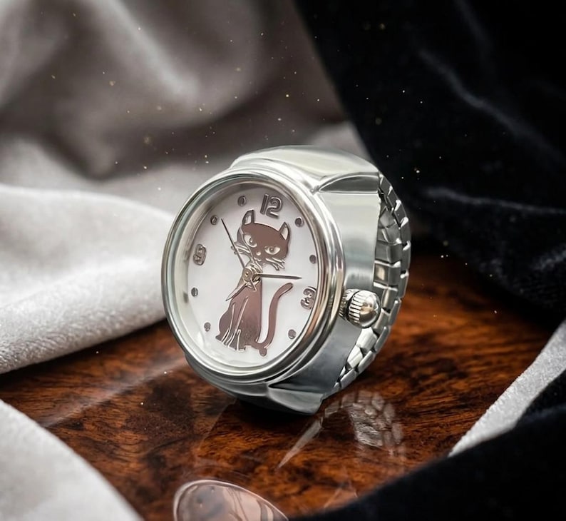 May include: A silver ring watch with a white face featuring a brown cat design. The watch has a round face with black numerals at the 12, 3, and 9 o'clock positions. The watch band is made of silver links.
