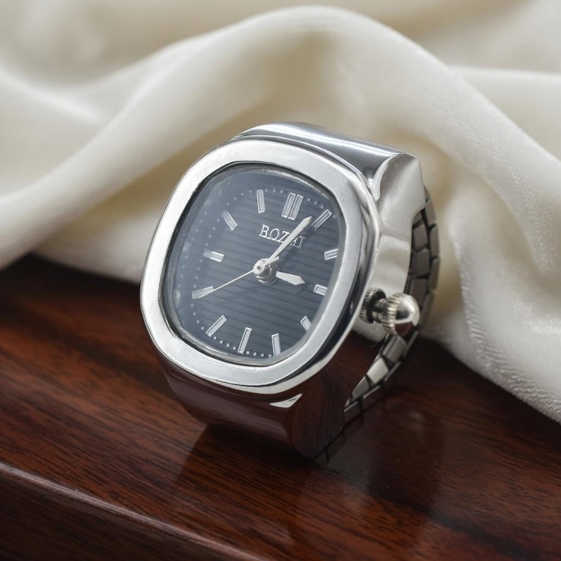 May include: A silver-toned ring watch with a dark gray face. The watch has a rectangular face with the word "BOZAI" printed on it. The watch band is a flexible, expandable design. The watch is displayed on a dark wooden surface.