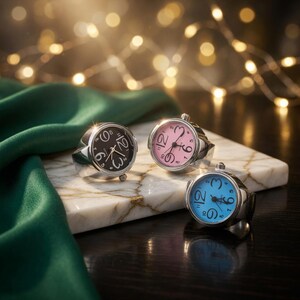 May include: Three silver-toned watch rings with black, pink, and blue watch faces. The rings are displayed on a marble surface with a green cloth and a string of lights in the background. Each ring has a different colored watch face.