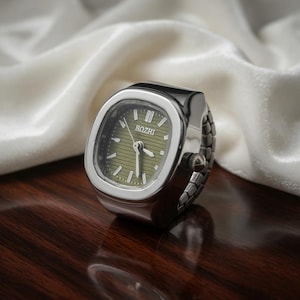 May include: A silver-toned ring watch with a rectangular face and a green dial. The watch face displays the brand name "BOZHI". The band is made of linked metal pieces. The watch is set on a dark wooden surface.
