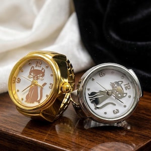May include: Two ring watches with white faces and cat designs. One watch has a gold band and a brown cat, the other has a silver band and a silver cat. Both watches have black hands and the numbers 12, 3, and 9.