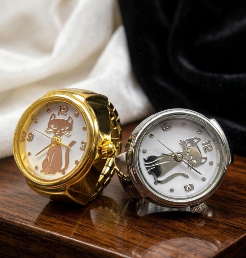 May include: Two ring watches with white faces and cat designs. One watch has a gold band and a brown cat, the other has a silver band and a silver cat. Both watches have black hands and the numbers 12, 3, and 9.