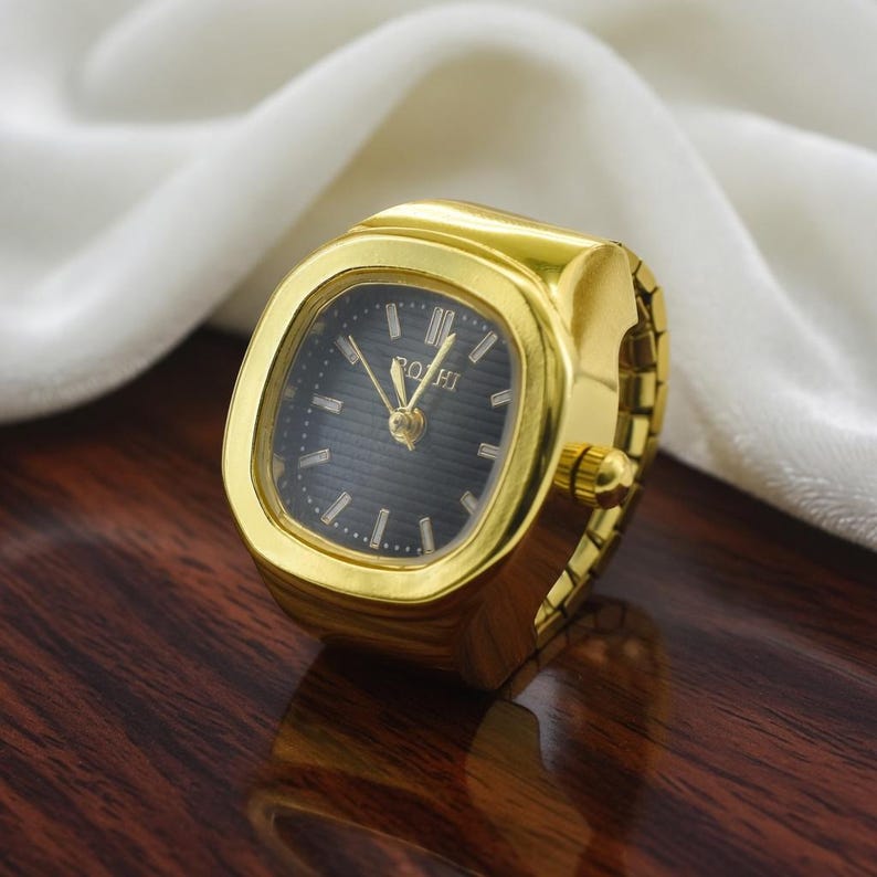 May include: A gold-toned ring watch with a square black face. The watch has a gold band and a small crown. The watch face has gold hands and the word "ROHT" printed on it. The watch is on a dark wooden surface.