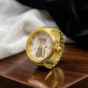 May include: A gold-toned ring watch with a white face featuring a cat silhouette and the number 12. The watch has a classic design with a round face and a band that resembles a watch bracelet. The ring is displayed on a wooden surface.