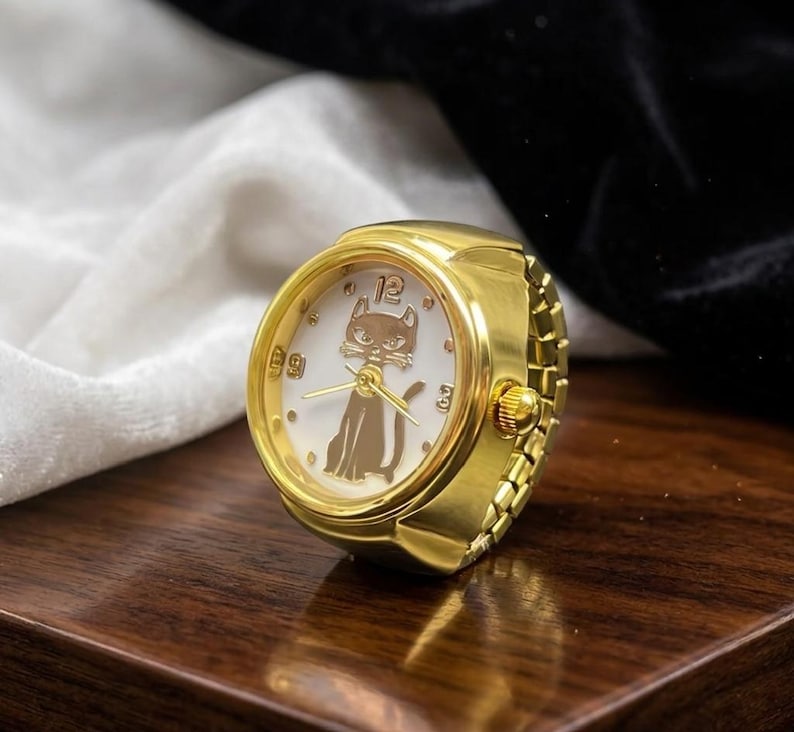 May include: A gold-toned ring watch with a white face featuring a cat silhouette and the number 12. The watch has a classic design with a round face and a band that resembles a watch bracelet. The ring is displayed on a wooden surface.