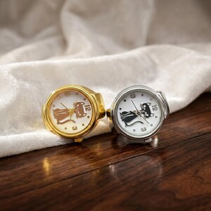 May include: Two ring watches with white faces and cat designs. One ring is gold-toned, the other is silver-toned. The watch faces display the numbers 3 and 12. The rings are set against a white cloth and a wooden surface.