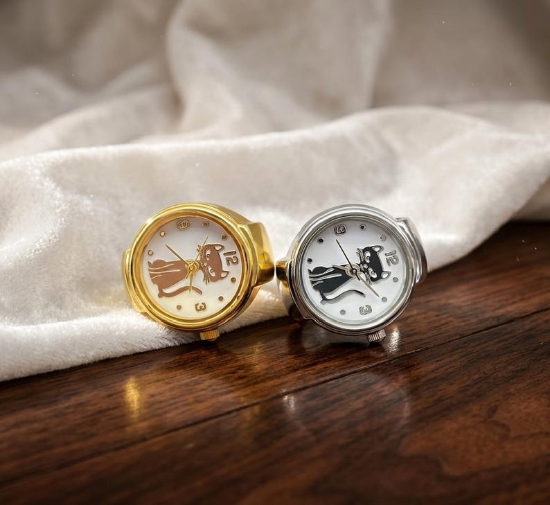 May include: Two ring watches with white faces and cat designs. One ring is gold-toned, the other is silver-toned. The watch faces display the numbers 3 and 12. The rings are set against a white cloth and a wooden surface.