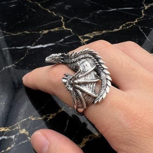 Op de afbeelding: Een zilveren drakenring met ingewikkelde details. De kop en staart van de draak omsluiten de vinger, met uitgespreide vleugels. Een accessoire met een fantasiethema.