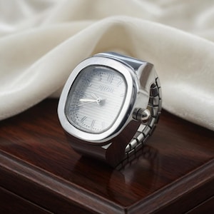May include: A silver ring watch with a rectangular face and a silver band. The watch face has Roman numerals and the brand name "BOZII". The watch is displayed on a dark wooden surface.