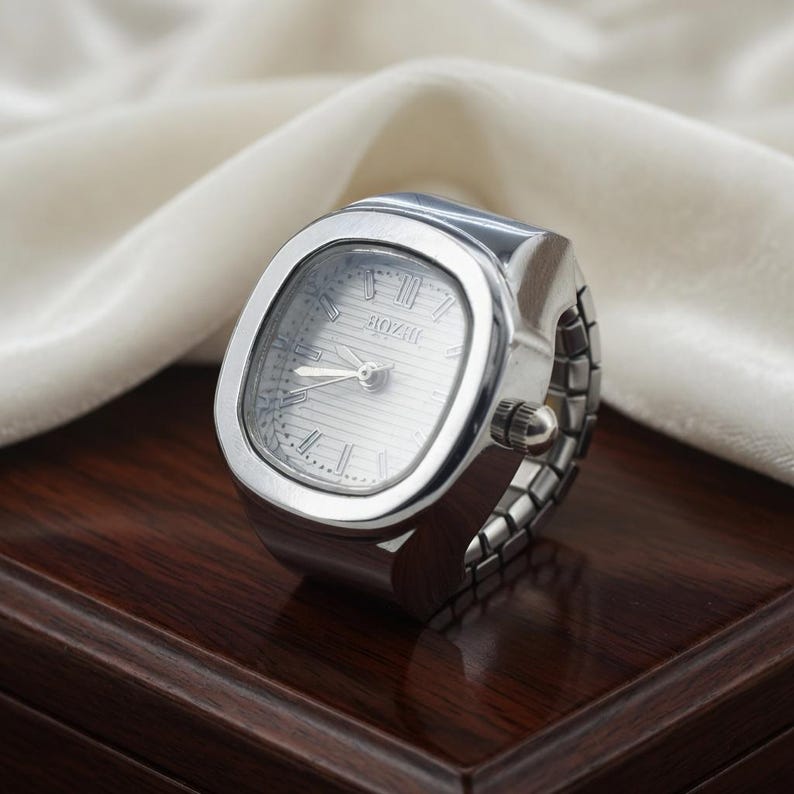 May include: A silver ring watch with a rectangular face and a silver band. The watch face has Roman numerals and the brand name "BOZII". The watch is displayed on a dark wooden surface.