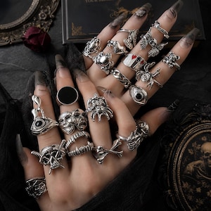 May include: A collection of silver-toned metal rings worn on fingers, featuring various designs including skulls, an eye with a pupil, a spider, playing card suits, masks, fangs, bones, and wings. The rings have a polished finish and intricate details.