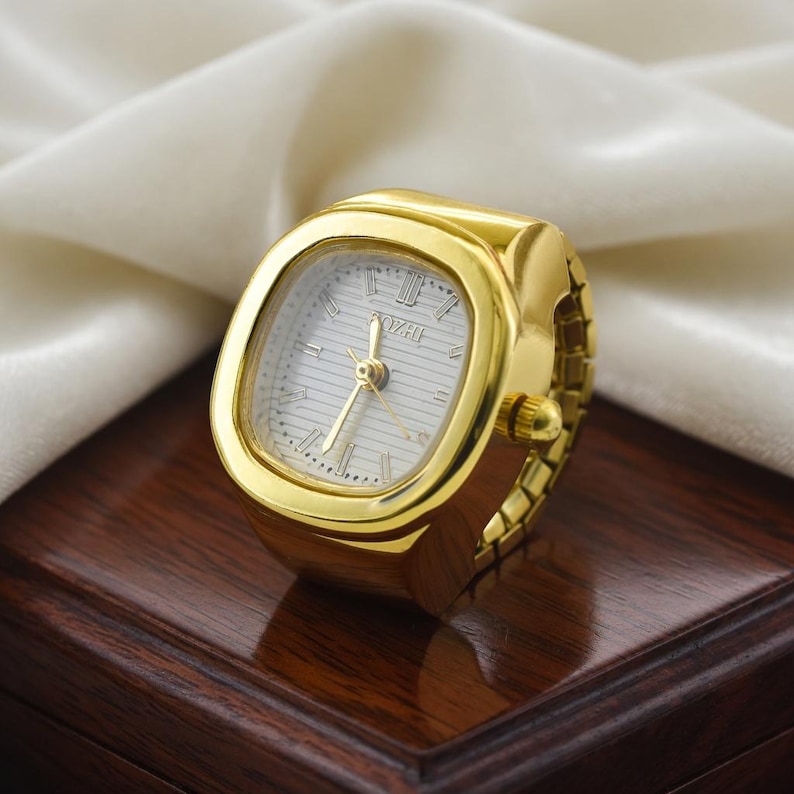 May include: A gold-toned ring watch with a rectangular face and Roman numeral hour markers. The watch face is white with gold hands and the band is an expandable style. The watch is displayed on a dark wooden surface.