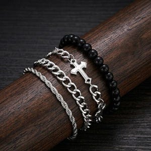 May include: Four bracelets displayed on a wooden surface. The bracelets include a black beaded bracelet, a silver chain bracelet with a cross pendant, a silver chain bracelet, and a silver rope bracelet. The bracelets are a mix of textures and styles.
