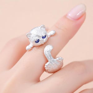 May include: A silver cat ring with blue eyes. The ring features a cat head on one side and a tail on the other, creating a wrap-around design. The cat's face has a slightly sad expression.