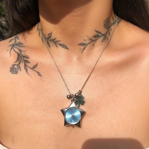 May include: Silver star-shaped pendant watch with a light blue face, suspended from a delicate silver chain. The pendant features a small charm and a four-leaf clover. The necklace is a piece of jewellery.
