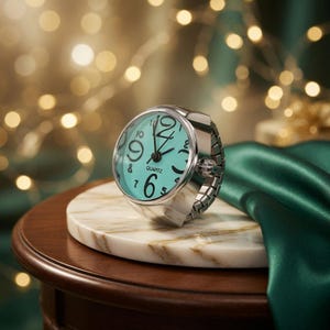 May include: A silver-toned ring watch with a turquoise face and black numerals. The watch is set on a white marble surface, with a dark wood table and green satin fabric in the background. The watch band is a silver-toned, expandable style.