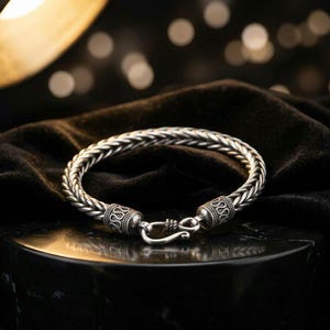 Puede incluir: Una pulsera de cadena plateada con un diseño de patrón de trigo trenzado. La pulsera incluye dos tapas cilíndricas en los extremos con detalles de volutas grabadas y un mecanismo de cierre de gancho, con un acabado metálico pulido.