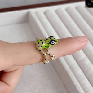 Puede incluir: Un anillo de rana caprichoso. El anillo presenta un diseño de rana con lunares verdes y negros con un cuerpo bronceado y patas doradas. El ojo de la rana es negro con un reflejo blanco. El anillo se lleva en un dedo.