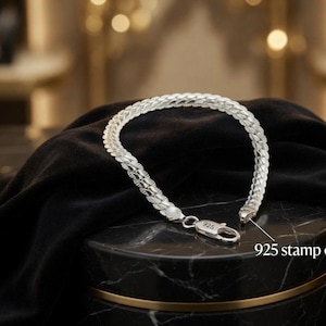 Puede incluir: Una pulsera de cadena plateada con un diseño de eslabones entrelazados y texturizados. La pulsera incluye un cierre de mosquetón con un sello 925 claramente visible, que indica una composición de plata esterlina. El metal tiene un acabado brillante.