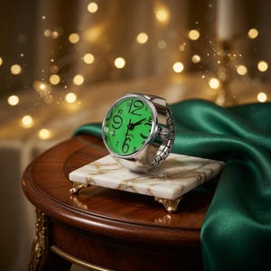 May include: A silver ring watch with a green face displaying the time. The watch sits on a marble and gold-trimmed stand. The background features a dark wooden table, green fabric, and bokeh lights.