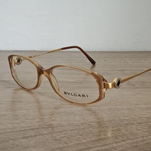 Könnte beinhalten: Eine Bvlgari-Brille mit hellbraunem Rahmen und goldfarbenen Bügeln. Die Gläser sind oval. Der Markenname ist auf dem Rahmen aufgedruckt. Die Bügel haben eine dunkelbraune Spitze. Auf einer Holzoberfläche.