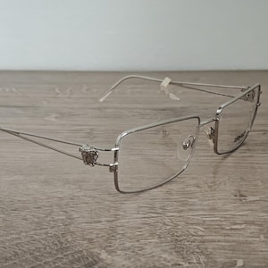 May include: Silver-framed eyeglasses with rectangular lenses. The frames feature a decorative emblem on the temples. The glasses are set on a wooden surface. The lenses are clear and the arms are thin.