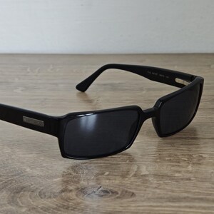 May include: Black rectangular sunglasses with dark lenses. The sunglasses have a sleek, modern design with a silver accent on the temples. The frames are made of a glossy black material, and the arms curve slightly.