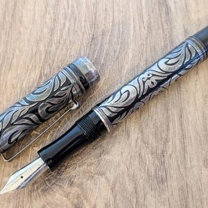 以下が含まれることがあります： 銀色の花柄彫刻と銀色のペン先が付いた黒い万年筆。 ペンのキャップは外されており、ペン先とバレル上の複雑なデザインが明らかになっています。 ペンは木製の表面に置かれています。