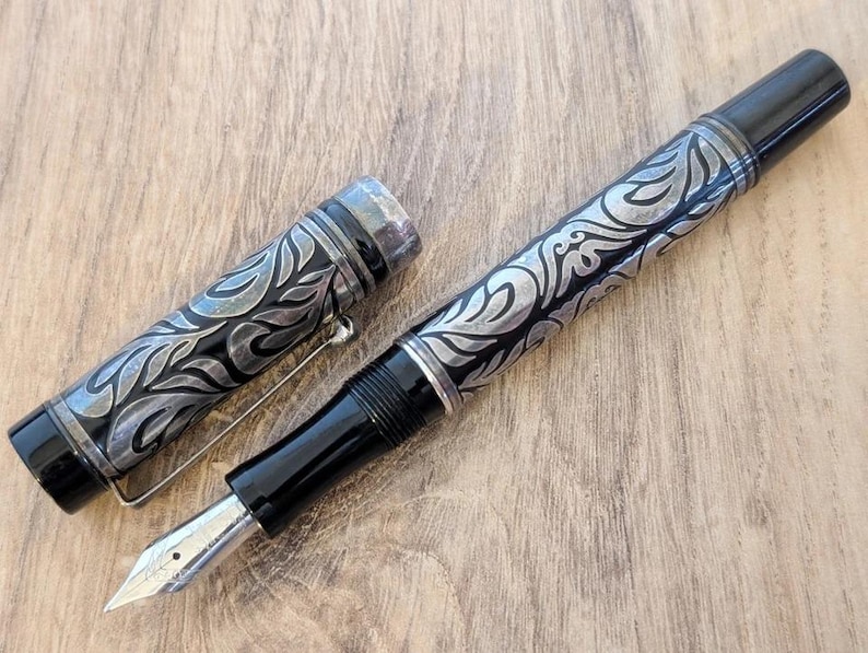 以下が含まれることがあります： 銀色の花柄彫刻と銀色のペン先が付いた黒い万年筆。 ペンのキャップは外されており、ペン先とバレル上の複雑なデザインが明らかになっています。 ペンは木製の表面に置かれています。
