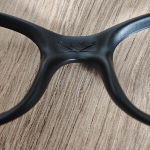 May include: Close-up of black eyeglasses. The frame is matte black with a subtle logo detail at the bridge. The arms curve smoothly, and the overall design is sleek and modern. The glasses are set against a neutral, textured background.