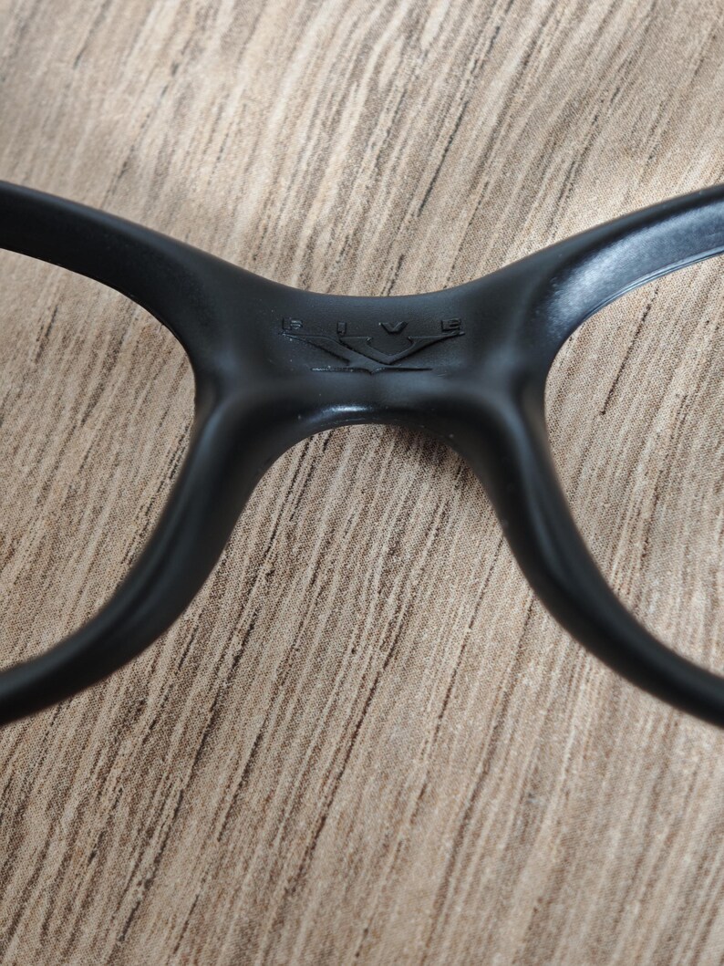May include: Close-up of black eyeglasses. The frame is matte black with a subtle logo detail at the bridge. The arms curve smoothly, and the overall design is sleek and modern. The glasses are set against a neutral, textured background.