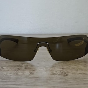 May include: Dark brown rimless sunglasses with a curved design. The sunglasses have a single bridge and dark lenses. The frame is a dark brown color. The sunglasses are resting on a wooden surface.
