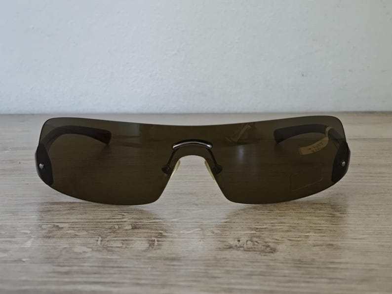 May include: Dark brown rimless sunglasses with a curved design. The sunglasses have a single bridge and dark lenses. The frame is a dark brown color. The sunglasses are resting on a wooden surface.