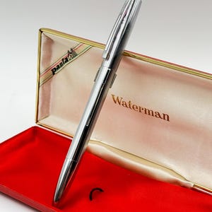 K&ouml;nnte beinhalten: Ein silberner Waterman-Stift mit gerilltem Design, pr&auml;sentiert in einem offenen roten und goldenen Stiftetui. Das Etui hat ein cremefarbenes Innere mit dem Waterman-Logo in goldfarbenen Buchstaben. Der Stift ist angewinkelt und zeigt seine elegante, metallische Oberfl&auml;che.
