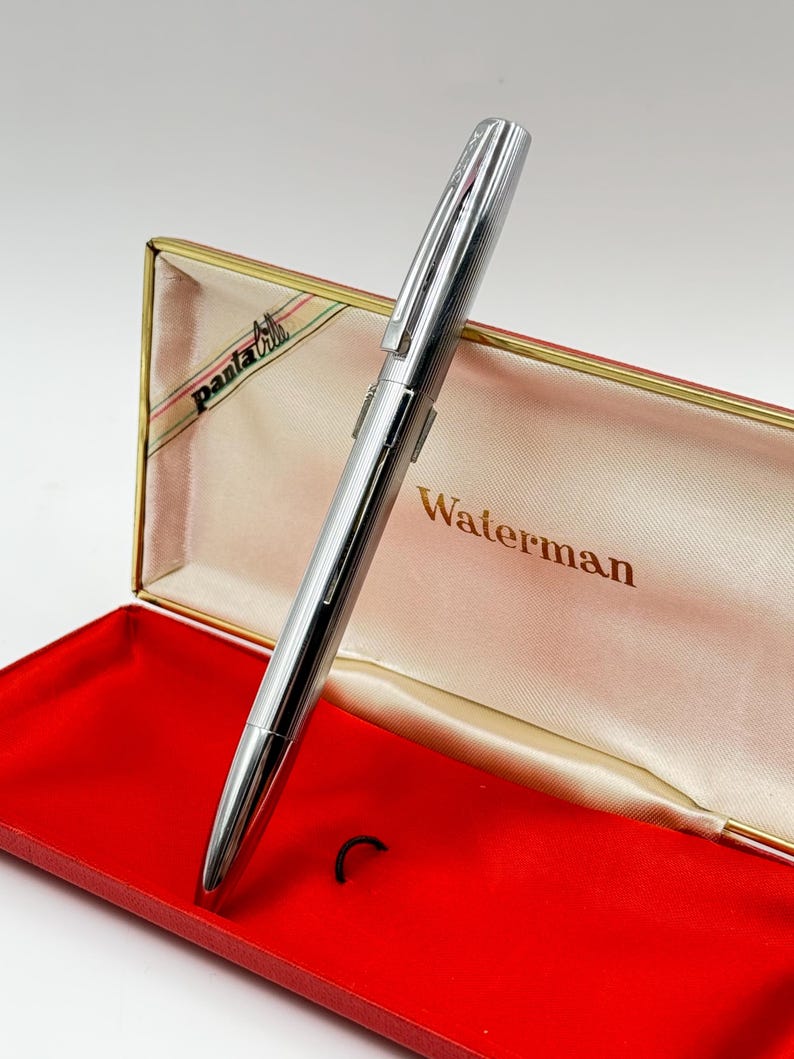 K&ouml;nnte beinhalten: Ein silberner Waterman-Stift mit gerilltem Design, pr&auml;sentiert in einem offenen roten und goldenen Stiftetui. Das Etui hat ein cremefarbenes Innere mit dem Waterman-Logo in goldfarbenen Buchstaben. Der Stift ist angewinkelt und zeigt seine elegante, metallische Oberfl&auml;che.