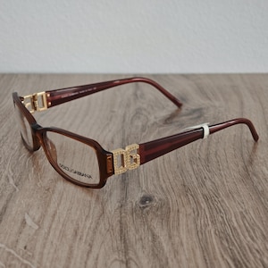 Puede incluir: Gafas Dolce & Gabbana marrones con montura rectangular. Las patillas son de un marrón más oscuro, y las bisagras y el logotipo DG son dorados. Las lentes son transparentes y el nombre de la marca está impreso en la lente izquierda.
