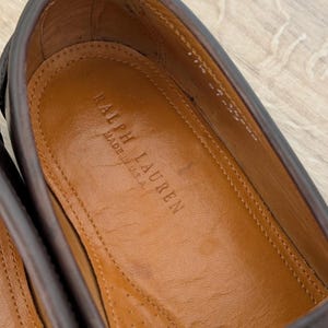 May include: Close-up of a brown leather shoe interior. The insole is tan with the embossed text "RALPH LAUREN MADE IN U.S.A." The shoe's upper is a darker brown, and the stitching is visible.