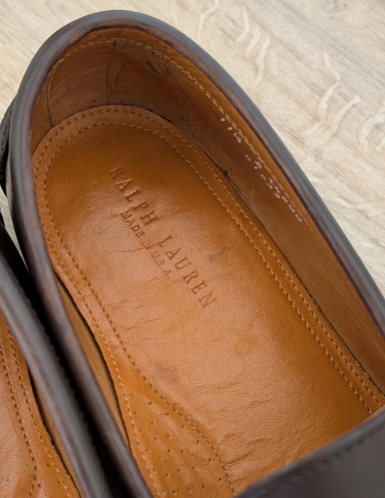 May include: Close-up of a brown leather shoe interior. The insole is tan with the embossed text "RALPH LAUREN MADE IN U.S.A." The shoe's upper is a darker brown, and the stitching is visible.