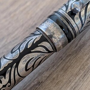 以下が含まれることがあります： 精巧な葉と花の模様が施された銀色のペンのクローズアップ。ペンには黒いバンドがあり、「Folli Follie」の文字が見えます。デザインは詳細でエレガントで、ヴィンテージ感があります。