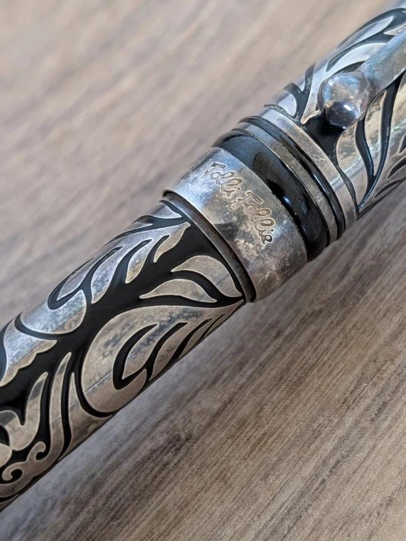 以下が含まれることがあります： 精巧な葉と花の模様が施された銀色のペンのクローズアップ。ペンには黒いバンドがあり、「Folli Follie」の文字が見えます。デザインは詳細でエレガントで、ヴィンテージ感があります。