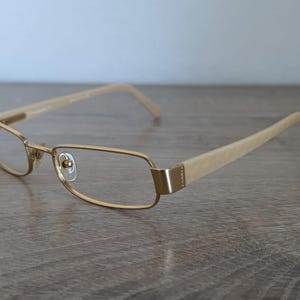 May include: Gold-tone eyeglasses with rectangular frames and light-colored temples. The glasses have a classic design, suitable for everyday wear. The lenses are clear, and the frames appear to be made of metal.