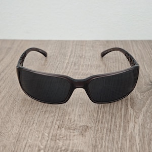 Puede incluir: Gafas de sol de color marr&oacute;n oscuro con un dise&ntilde;o de montura envolvente y lentes tintadas oscuras. Las patillas presentan un patr&oacute;n recortado distintivo en los laterales, lo que aporta un aspecto estructurado. La montura tiene un acabado liso.