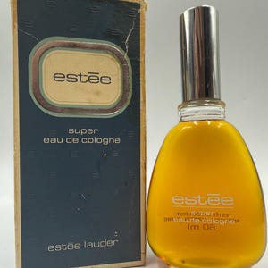 Op de afbeelding: Vintage Estée parfumfles en doos. De fles heeft een zilveren dop en is gevuld met gele vloeistof. De doos is donkerblauw met de naam "estée" in een gouden ovaal. De tekst "super eau de cologne" is zichtbaar.