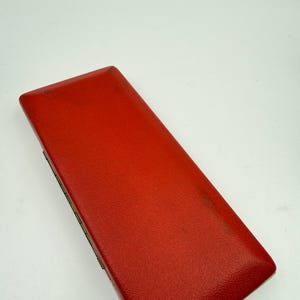 K&ouml;nnte beinhalten: Eine rechteckige, rote Box mit Lederstruktur. Die Box ist geschlossen und hat eine leicht abgerundete Oberseite. Die Kanten der Box sind dunkler rot. Die Box befindet sich vor einem wei&szlig;en Hintergrund.
